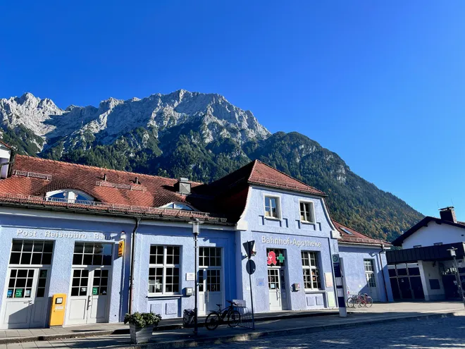 A pale blue station building with a red tiled roof beneath a limestone mountain ridge, under a cloudless sky