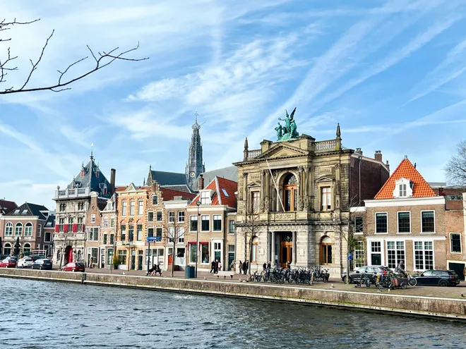 A row of canal-side buildings with a classical stone facade topped by a sculpture, a church spire behind, under a streaked blue sky
