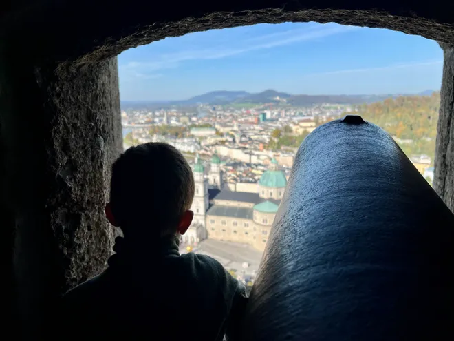 A boy looking out through an arched stone opening beside a cannon, the city below