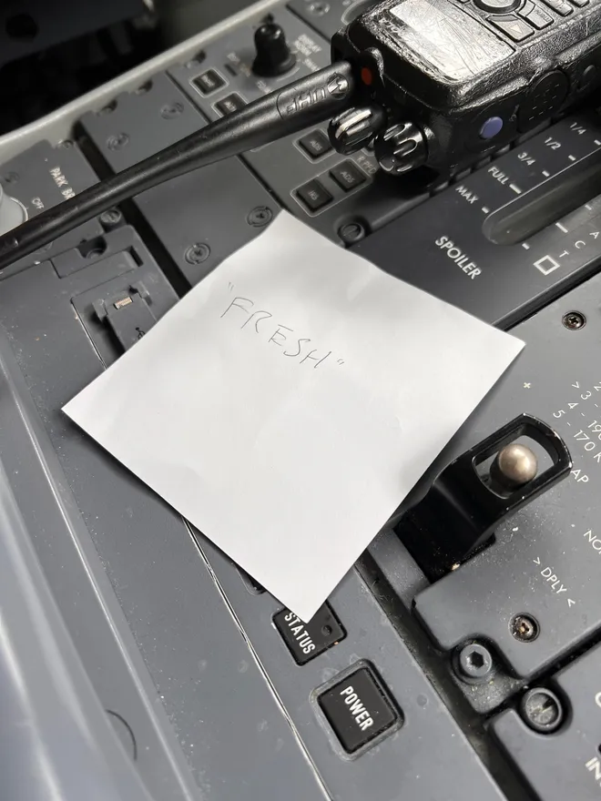 A handwritten note reading Fresh on a scrap of paper, lying on an aircraft cockpit control panel beside a handheld radio
