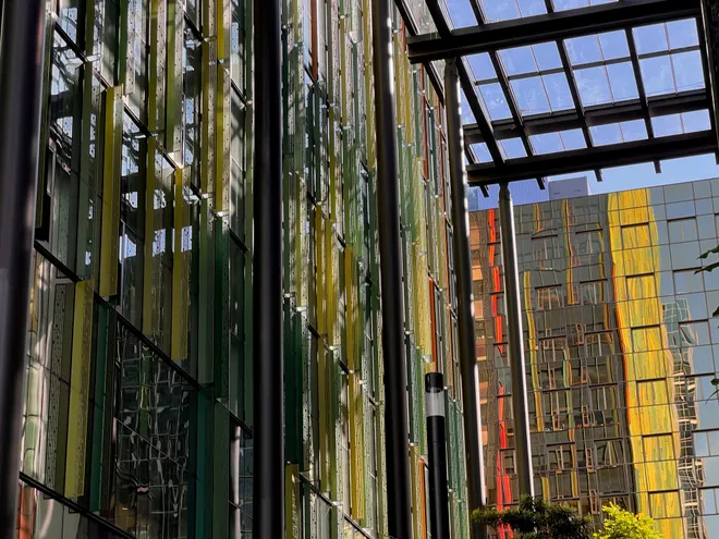 A wall of tall green and yellow glass panels under a steel canopy, a gold-tinted tower reflected beyond