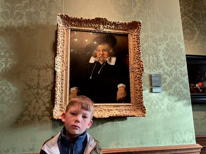 A boy standing beneath a framed Dutch portrait of a man in a broad hat and white collar, wearing much the same expression