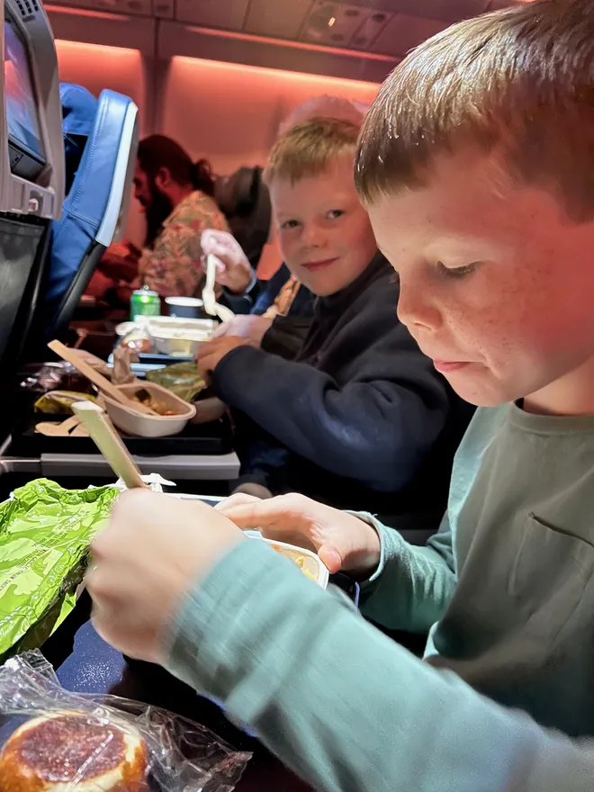 Two boys eating meals from trays in an aircraft cabin, one looking into the camera