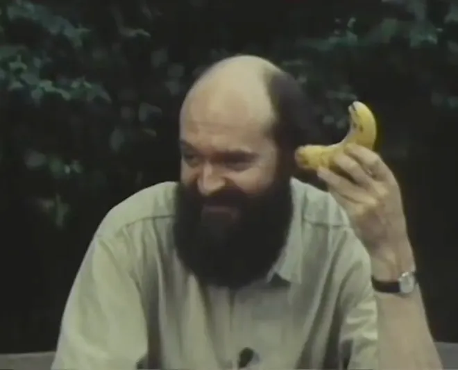 Arvo Pärt grinning, holding a banana to his ear like a telephone, dark foliage behind him