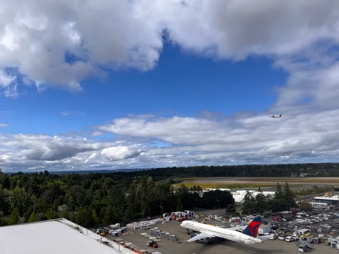 Looking down from a rooftop over an airport ramp, a parked Delta jet below and another climbing away above a line of evergreens