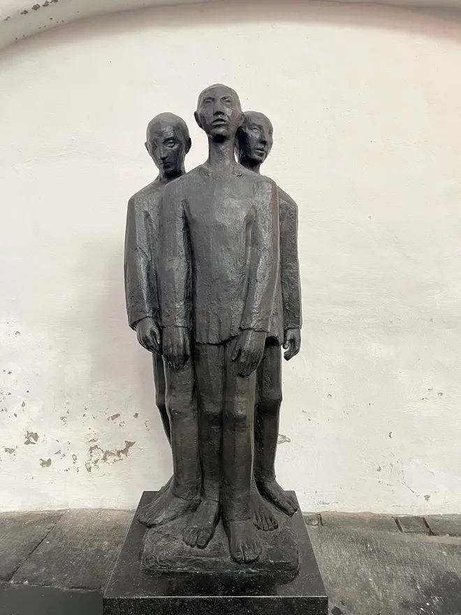 A dark bronze sculpture of three gaunt standing figures against a whitewashed wall