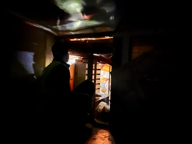 A person in a hi-vis vest crouched in a dark, cramped compartment, lit only by a glow from the equipment ahead