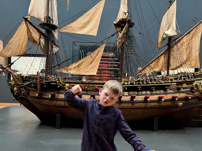 A boy flexing his arm in front of a large model of a tall ship in a museum hall